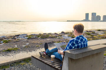 Man sitting on a bench by the sea and reading a newspaper with a takeaway coffee. Relaxing outdoors on a sunny day with a view of the city and coastline.