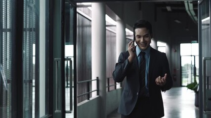 Professional asian businessman walking through modern office corridor, talking on smartphone with confident smile and engaging hand gestures while navigating corporate interior - Powered by Adobe