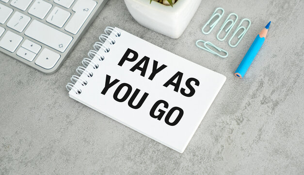 A white notebook with the words Pay As You Go written on it. A blue pencil is on the table next to the notebook