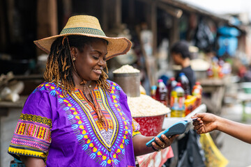 Contactless transactions, small business owner, SME, local traditional African market woman accepts payment via a mobile point of sale, POS system at an outdoor market stall.