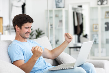 Man is sitting on a couch in his room with a laptop, feeling happy.