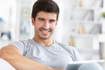 Handsome young man looking at camera close-up portrait in living room with electronic tablet