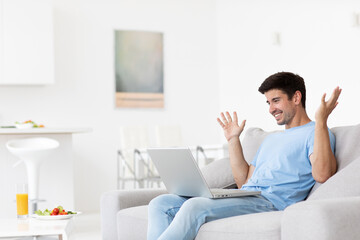 Man is sitting on a couch in his room with a laptop, feeling happy.