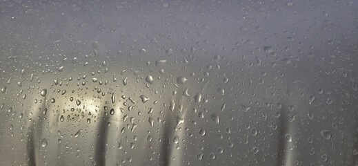 A fogged glass surface covered in water droplets reveals blurred vertical shapes behind it, possibly fence posts. The grey tones and condensation evoke a calm, rainy atmosphere