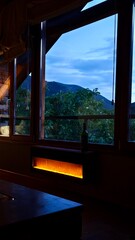 Fototapeta premium Large windows reveal a picturesque mountain view against a twilight sky. Inside, a warm glow emanates from a modern, wall-mounted electric fireplace. Romantic rustic relax atmosphere. 