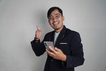 A happy Asian man gives a thumbs-up while holding a smartphone, conveying approval and confidence. This image is perfect for themes of business success, positive feedback, and modern technology