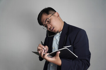 A busy Asian man in a blazer multitasks, talking on a phone while writing in a notebook. This image is perfect for themes of work, productivity, and professional stress.