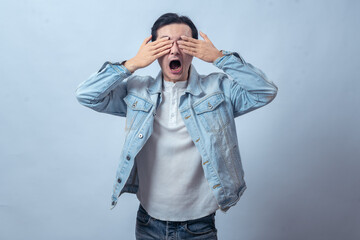Asian man in denim jacket covering part of his face with one hand, looking serious and thoughtful,...