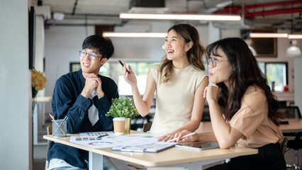 Asian woman designer holding pen smiling or laughing with coworkers at wooden table in the office.