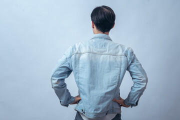 Asian man in denim jacket standing with hands on hips, seen from behind, posing in studio against plain light background, showing confident stance and casual modern fashion style