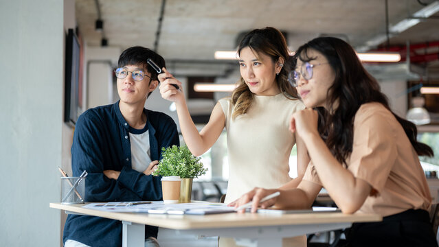 Asian woman designer holding pen pointing ahead for coworkers to look at wooden table in the office.