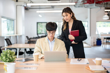Asian businesswoman looking at laptop supervising an intern coworker at wooden table in the office.