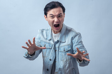 Asian man in denim jacket shouting with wide open mouth and raising hand forward, standing in...