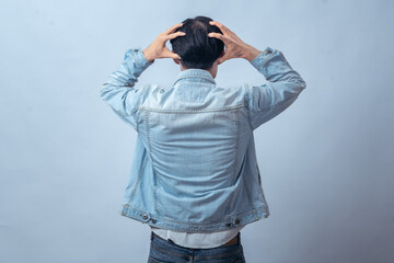 Asian man in denim jacket seen from behind, holding head with both hands in frustration, standing in studio against plain light background, expressing stress, tension, and emotional struggle