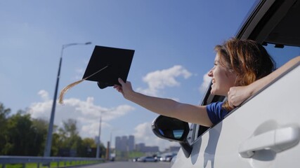 A Caucasian woman waves her graduation cap while extending her arms out of a car window.