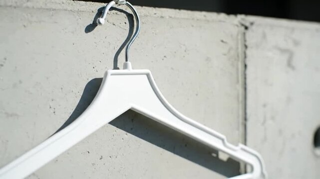 Close up of a modern white plastic clothes hanger featuring a reflective metal hook casting a sharp shadow on a light grey textured concrete wall in bright sunl
