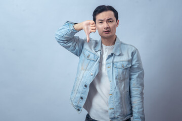 Asian man in denim jacket showing two thumbs down with a displeased expression, expressing dislike...