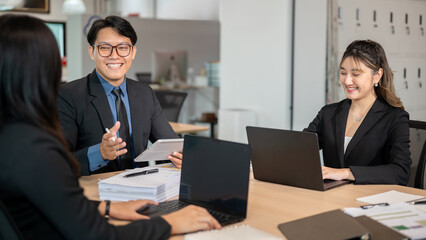 Smiling businessman holding tablet talking to woman coworker while sitting at wooden table in office