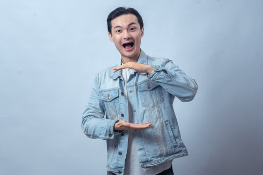 Asian man in denim jacket smiling and making space between hands as if holding invisible object against plain studio background, showing product presentation or measurement gesture in lifestyle pose