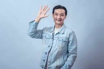 Smiling Asian man in denim jacket raising hand with open palm waving gesture against plain studio...