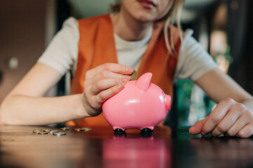 young European woman is sitting at her home table, carefully dropping coins into a ceramic piggy bank