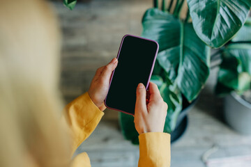 The hands of a young European woman, dressed in the sleeve of a vibrant yellow blazer, are holding a smartphone.