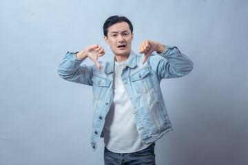 Asian man in denim jacket showing two thumbs down with a displeased expression, expressing dislike...