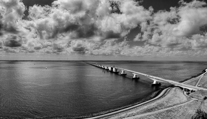 A long bridge, the Zeeland Bridge, stretches across a calm expanse of water. The sky is filled with dramatic clouds, casting shadows on the landscape.