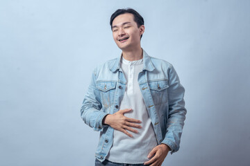 Asian man in denim jacket touching stomach with pained face, standing in studio against plain light background, expressing stomachache, discomfort, and sick feeling with casual fashion style