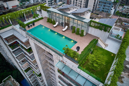 High-angle aerial view of a modern rooftop swimming pool with a wooden deck, lounge chairs, and solar panels, set against an urban city backdrop. Perfect for themes of architecture, urban lifestyle