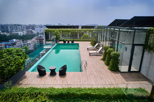 Modern rooftop swimming pool captured from above, featuring a wooden deck, lounge seating, and solar panels against an urban backdrop. Suitable for architecture, real estate, and lifestyle themes.