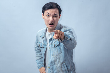 Asian man in denim jacket pointing finger directly at camera with serious face, standing in studio against plain light background, showing accusation, warning, and commanding gesture
