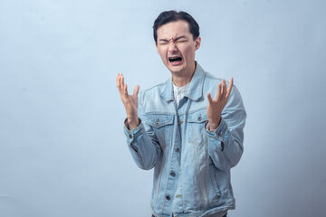 Asian man in denim jacket screaming with both hands raised, expressing anger, frustration, sadness,...