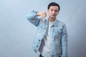 Asian man in denim jacket showing two thumbs down with a displeased expression, expressing dislike...