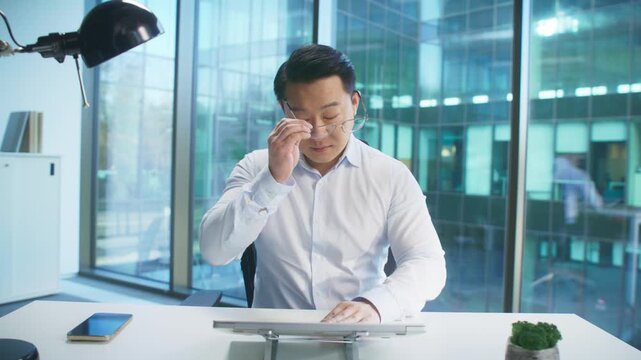 Calm Asian male removing glasses while closing laptop in stylish office. Wrapping up work or ending meeting. Pausing for break or clearing thoughts before leaving desk. Preparing to unwind.