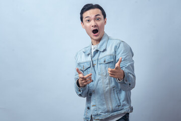 Asian man in denim jacket smiling and pointing with finger guns gesture against plain background, symbolizing fun, confidence, casual lifestyle, and positive youthful energy