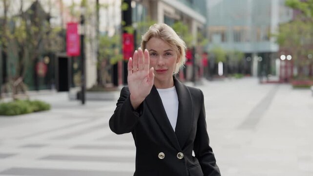 Portrait of woman business leader shows Stop gesture near modern office buildings. Blonde lady entrepreneur raises arm with open palm in ban gesture standing on sunny city street