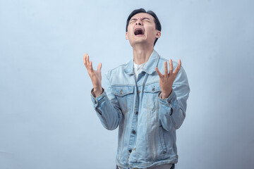 Naklejka premium Asian man in denim jacket screaming with both hands raised, expressing anger, frustration, sadness, or despair on a plain studio background, dramatic emotional reaction
