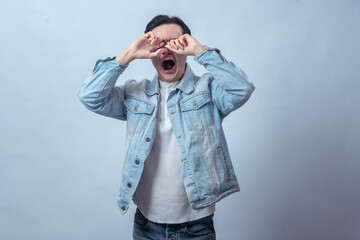 Asian man in denim jacket yawning with eyes closed and hand rubbing face, looking tired and sleepy, standing in studio against plain light background, expressing fatigue and exhaustion