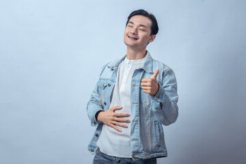 Asian man in denim jacket smiling with eyes closed, touching stomach with one hand and showing thumbs up, standing in studio against plain light background, expressing relief and satisfaction