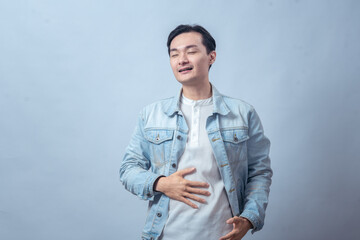 Asian man in denim jacket touching stomach with pained face, standing in studio against plain light background, expressing stomachache, discomfort, and sick feeling with casual fashion style