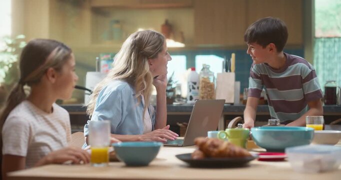 Mother Successfully Works on Remote Work on Her Laptop While Preparing a Wholesome Breakfast of Orange Juice, Cereal, and Croissants, Engaging With Her Joyful Children