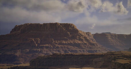 A stunning mountain range rises prominently against a backdrop of soft, swirling clouds and warm sunlight in the late afternoon. The rugged terrain showcases layers of rock formations.