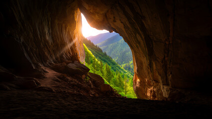 A view from inside a dark cave looking out at a lush green valley with mountains in the distance illuminated by sunlight