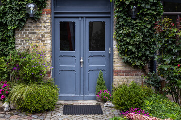 Blue Double Door Is Set In A Brick Wall Covered In Ivy And Surrounded By Lush Green And Purple...