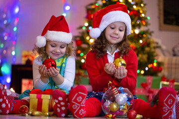 Little girls dressed as Santa Claus playing with gifts and Christmas tree decorations at home on Christmas.