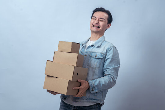 Asian man in denim jacket struggling to carry stack of cardboard boxes with tired expression against plain studio background, symbolizing heavy workload, delivery, shipping, or ecommerce service