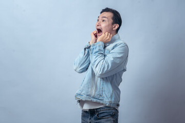Asian man in denim jacket showing scared expression, raising hands near face with open mouth in...