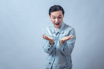 Asian man in denim jacket with shocked expression looking at his open palms, symbolizing surprise,...