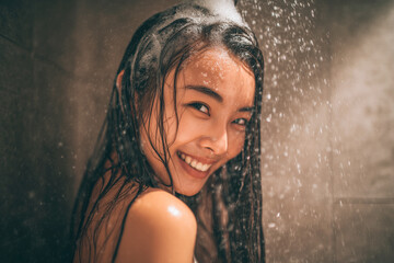 Fototapeta premium Portrait of a smiling asian woman with long wet hair, shampoo foam covering her head, standing in the shower